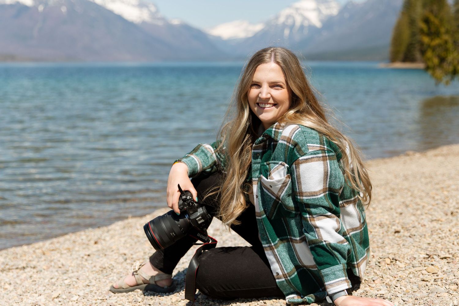 Photographer in plaid jacket sits by lake shore with mountains and blue water in background.