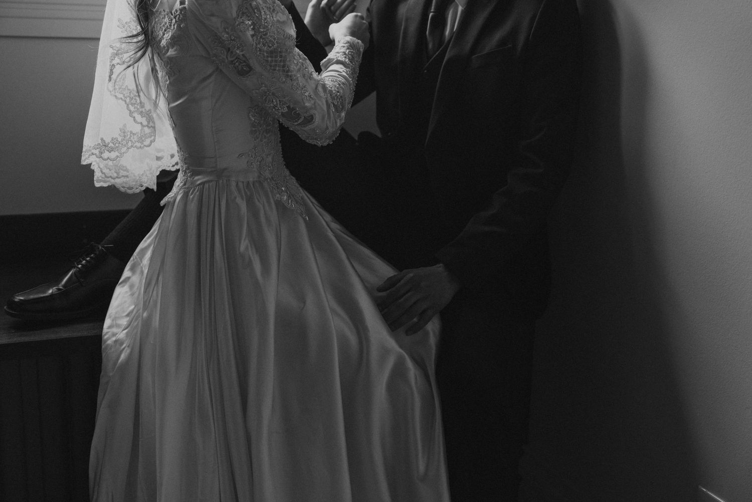 A black and white photograph shows a formal dress detail with delicate lace and flowing fabric in dramatic lighting.