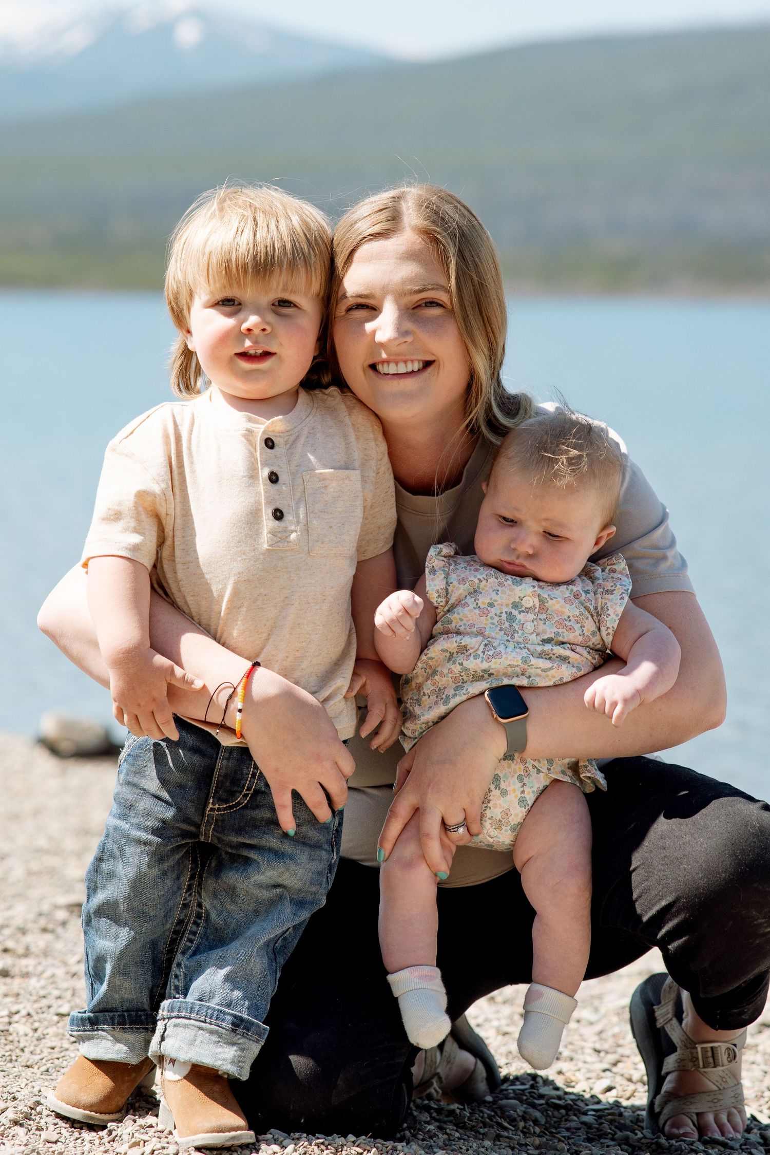 A family sits together outdoors by a lake with mountains in the background on a sunny day.