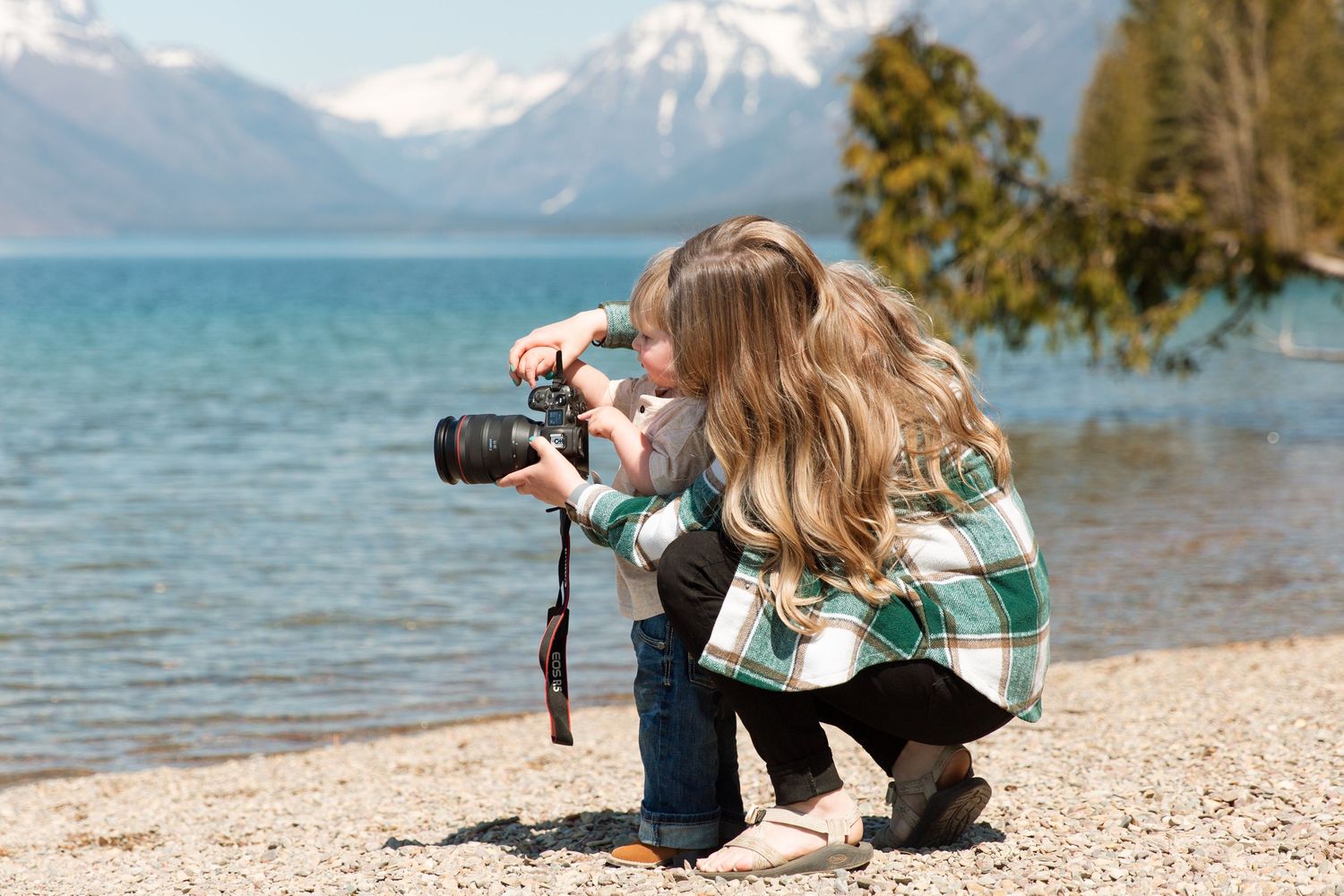 A photographer with a professional camera crouches at the shoreline of a turquoise lake with snow-capped mountains in the distance.