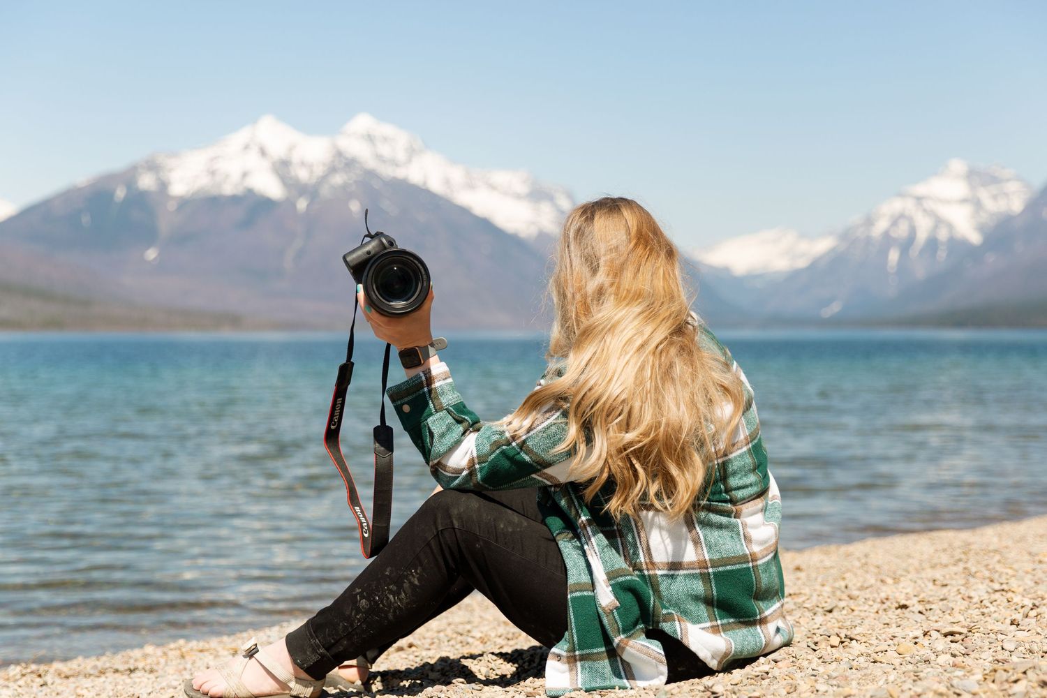Photographer with long blonde hair sits by turquoise lake with snowy mountains in background wearing plaid shirt.