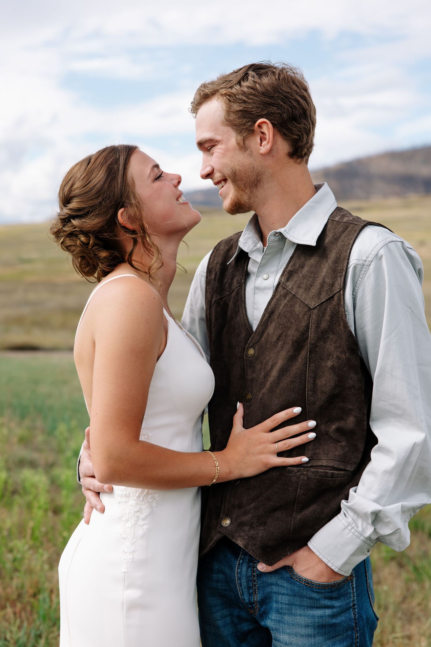 A couple in wedding attire share an intimate moment in a rustic outdoor setting with mountains in the background.