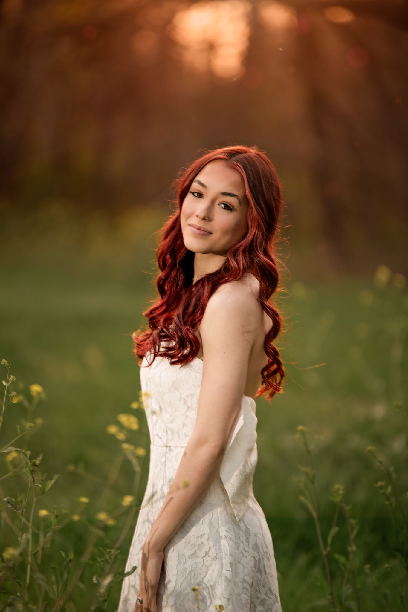A young woman in a white dress poses in a field during golden hour with flowing red hair.