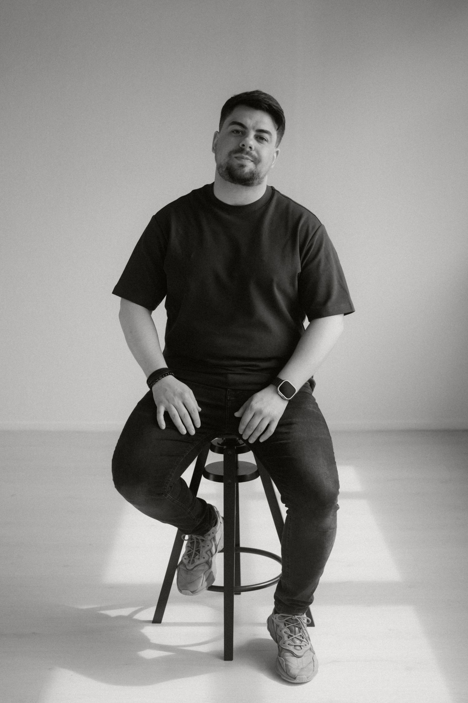 Black and white studio portrait of someone sitting casually on a bar stool wearing a t-shirt and sneakers.
