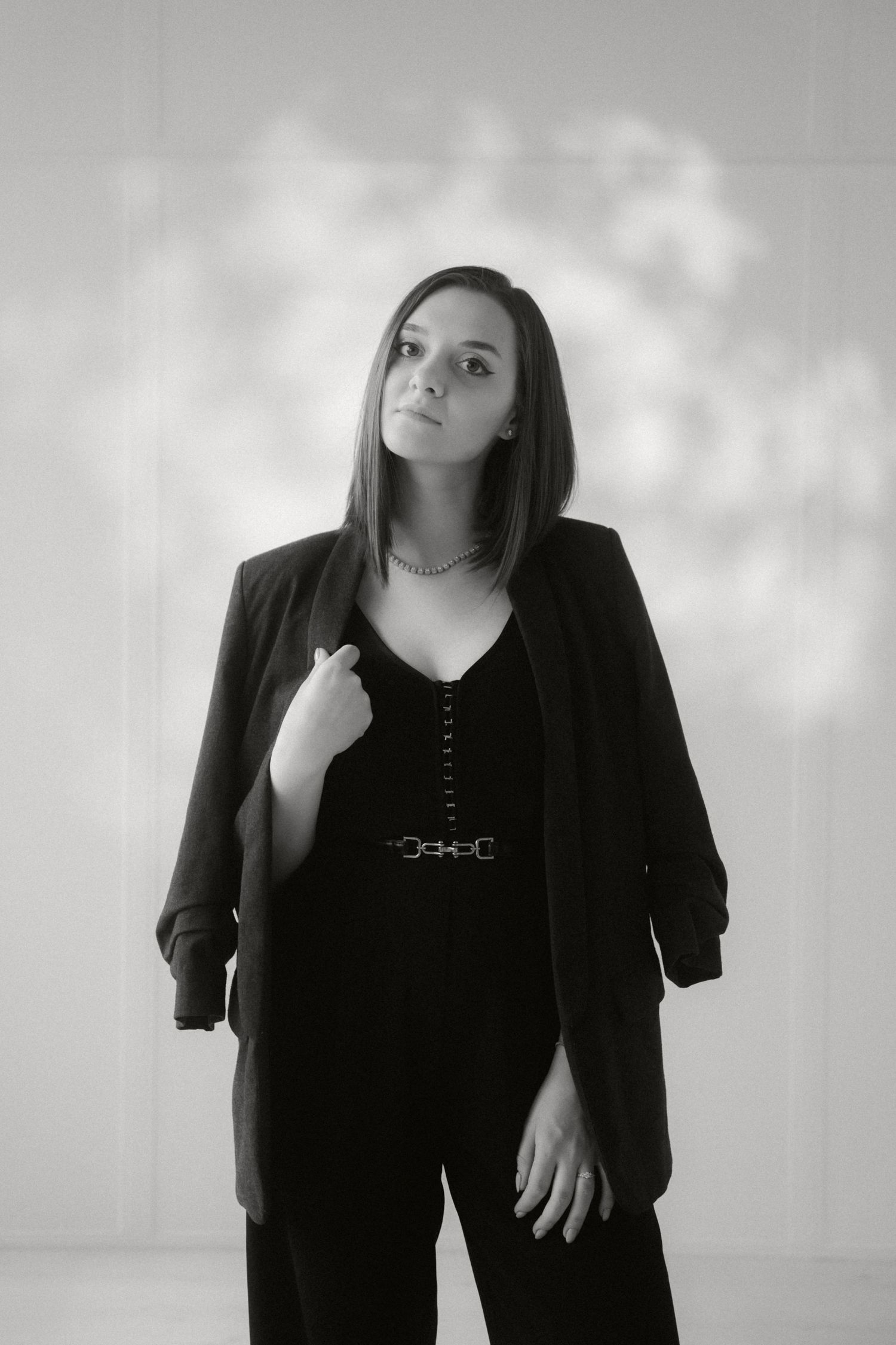 Black and white portrait of person in dark blazer and pants against light background with shadow pattern.