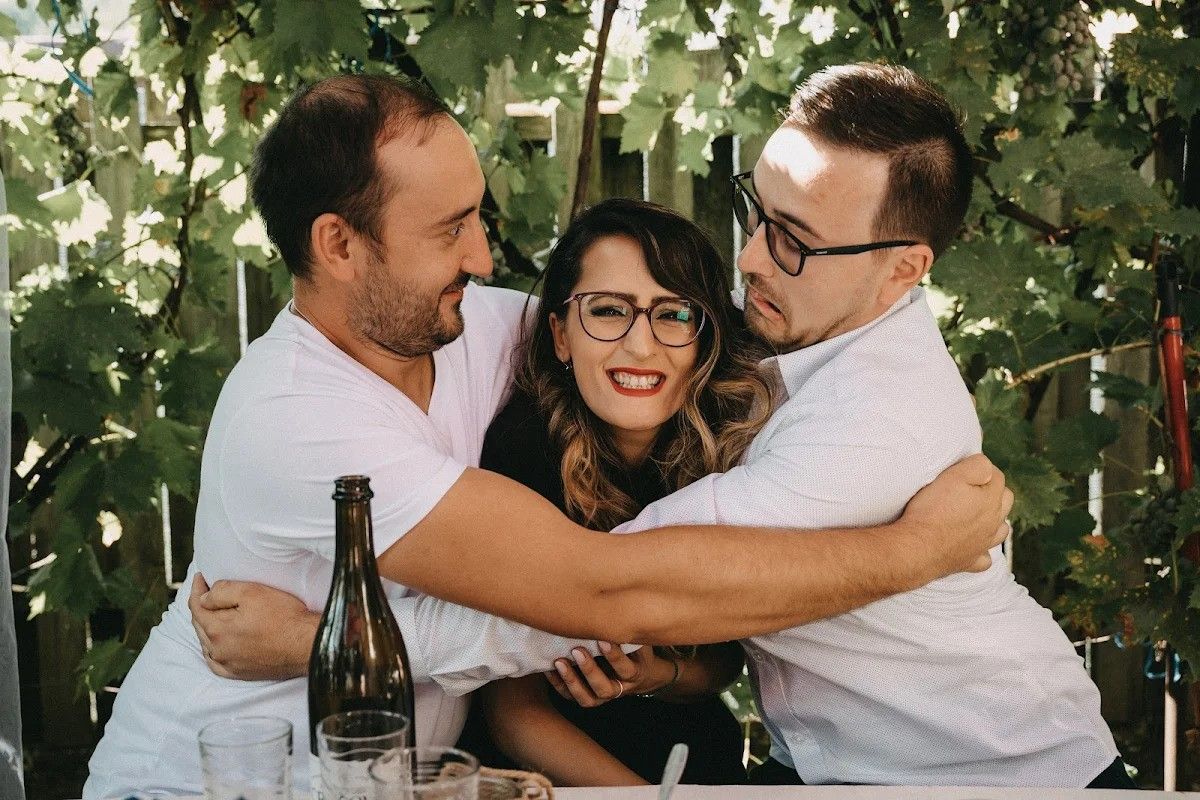 Three friends embracing in front of a table filled with wine bottles, sharing warmth and joy.