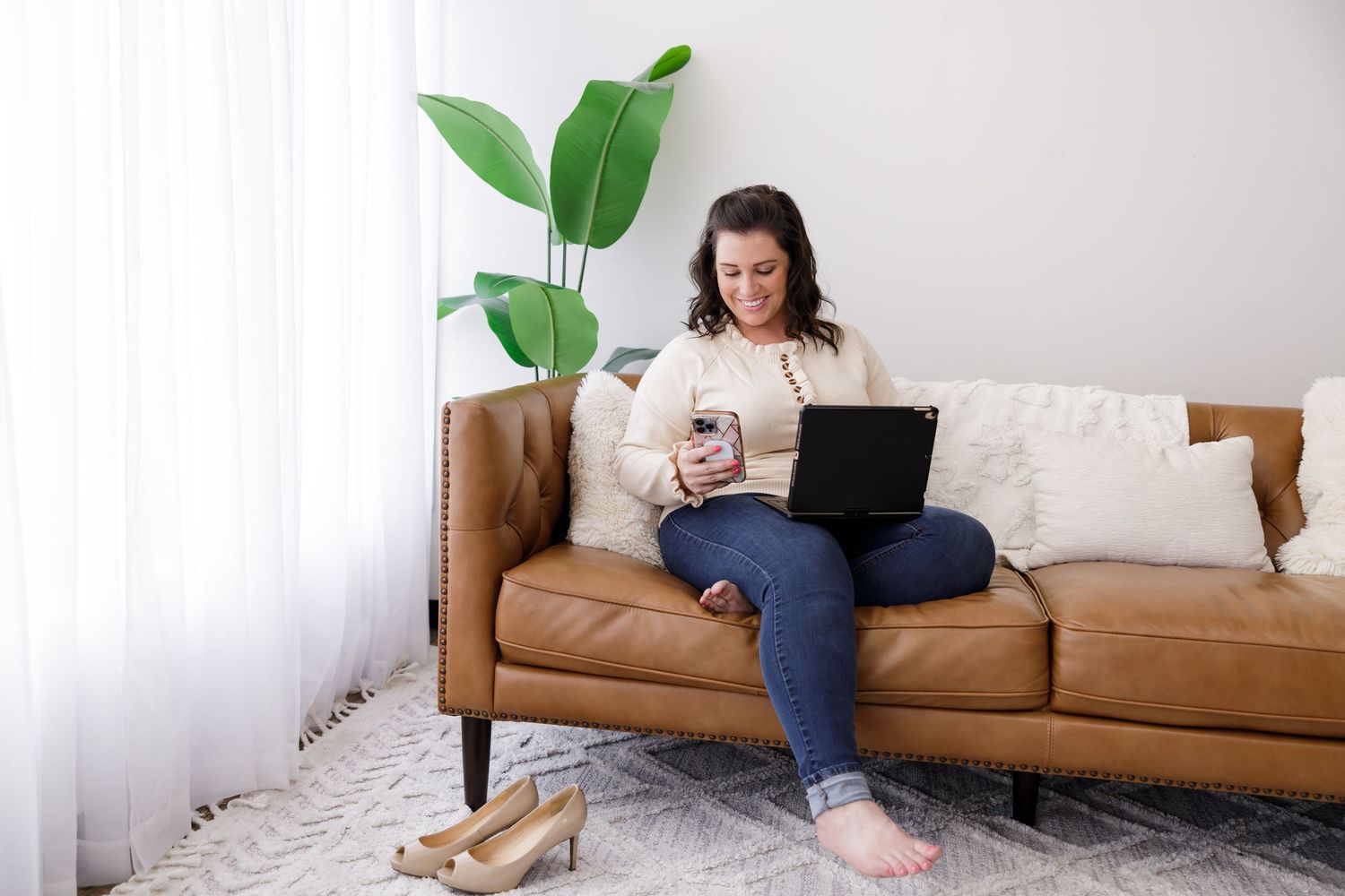 woman business owner sitting on couch in living room for lifestyle branding photo in Bothell, WA barefoot with phone in hand and laptop