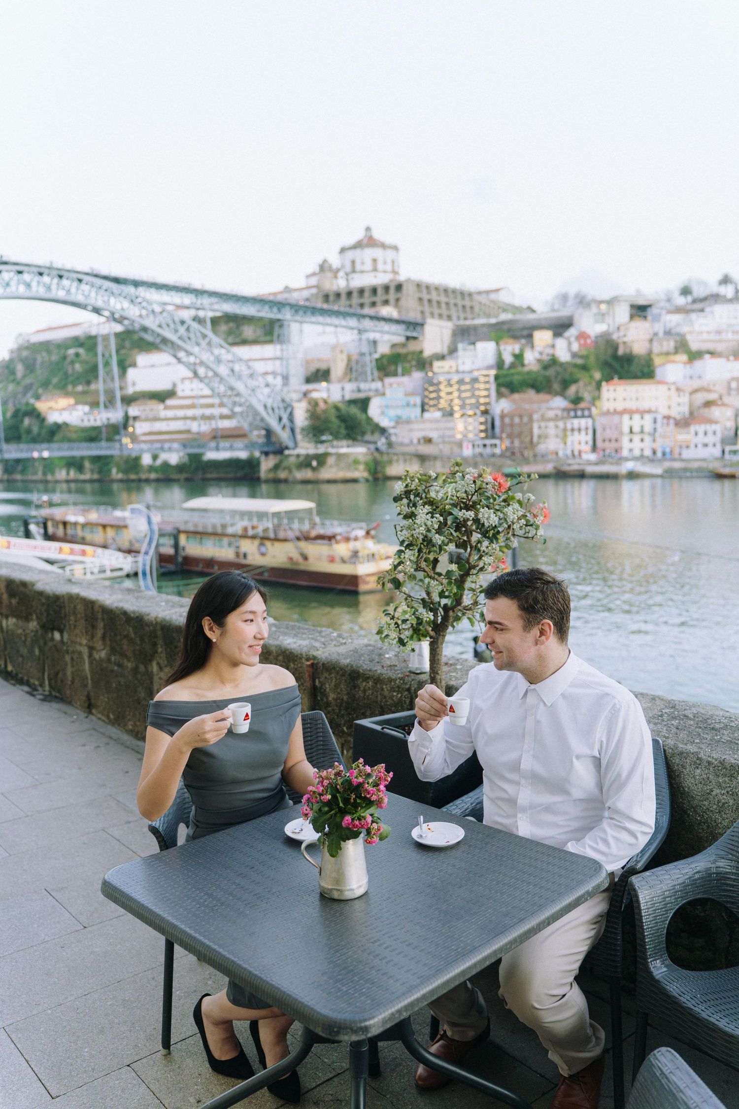A couple enjoys drinks at an outdoor cafe along the Douro River in Porto, with the iconic Dom Luis Bridge in the background.
