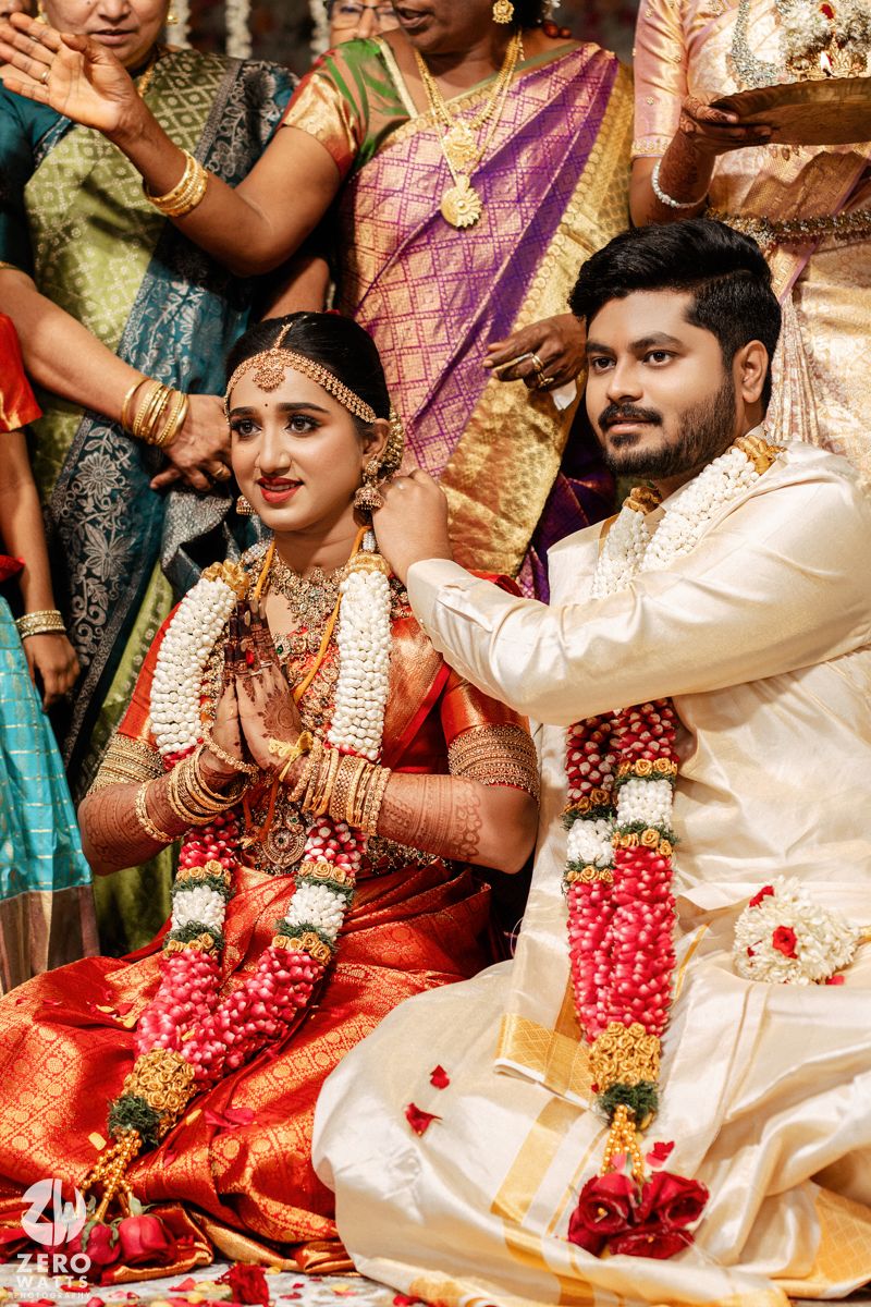 Traditional South Indian wedding ceremony with bride in red silk saree and groom in white attire wearing floral garlands.