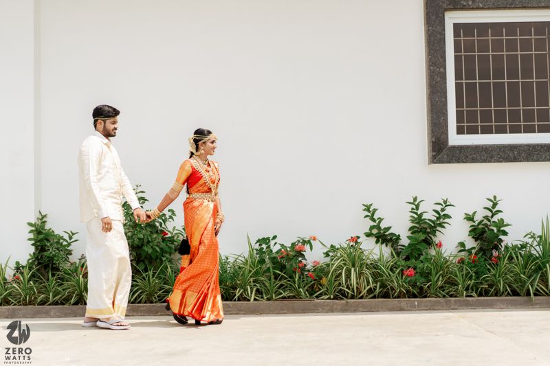A couple in traditional attire walking hand in hand along a white wall with green plants and flowers in the background.