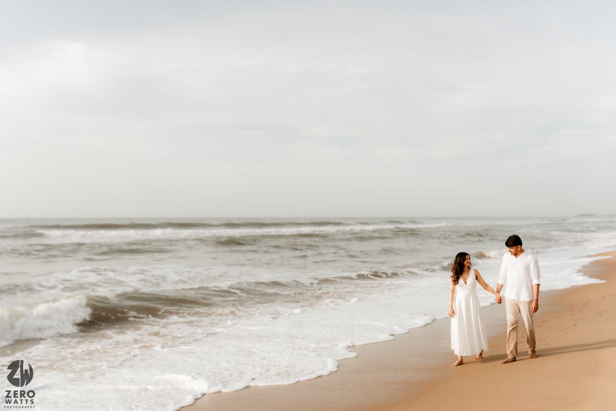 Two people in white walking hand in hand along a sandy beach with crashing waves.