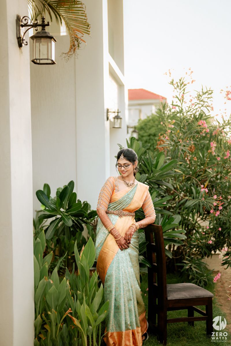 A person in a peach-colored sari stands in a white columned walkway surrounded by tropical plants and greenery.