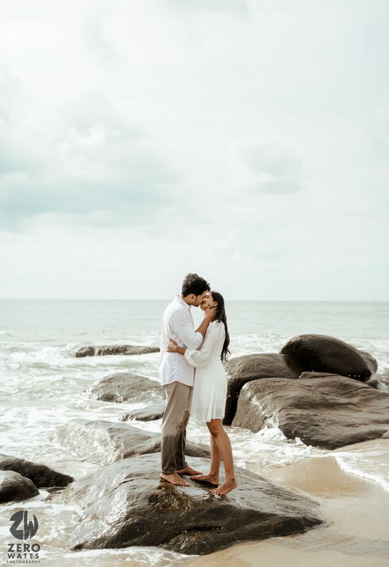 A romantic couple embraces on rocky coastal rocks as ocean waves crash around them on a cloudy day.