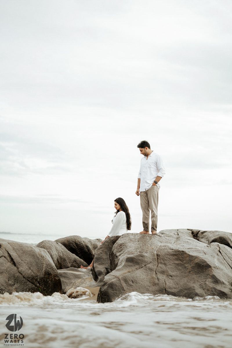 A couple stands together on rocky coastal terrain against an overcast sky and ocean waves.