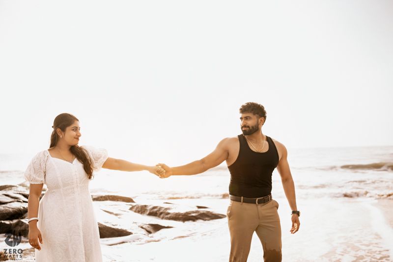 Couple holds hands while walking along rocky beach at sunset.