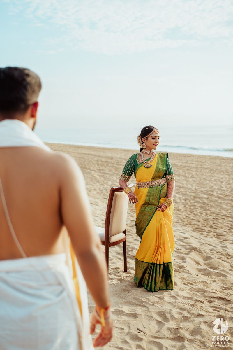 A couple in traditional South Asian attire pose for a romantic beach photoshoot with a wooden chair on sandy shores.