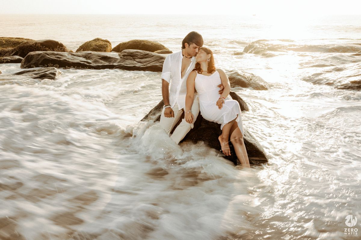 Couple in white outfits sitting on ocean rocks as waves splash around them.