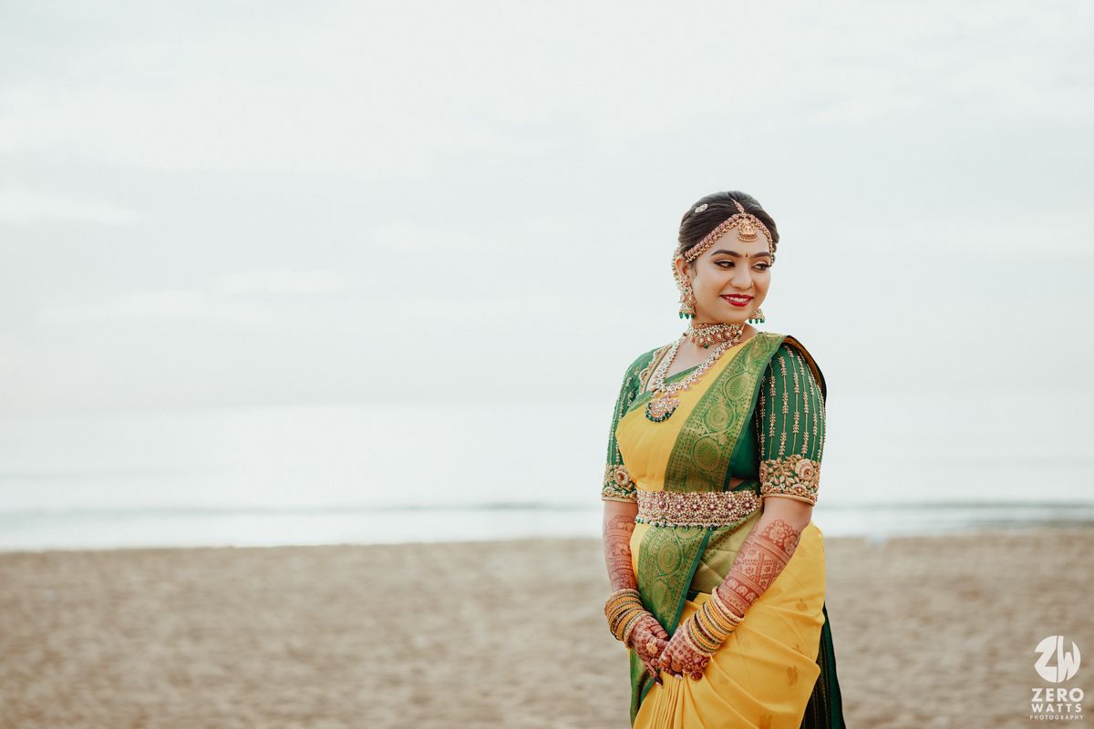Traditional South Indian Kanjeevaram silk saree in yellow and green with gold jewelry styled for a beachside photo.