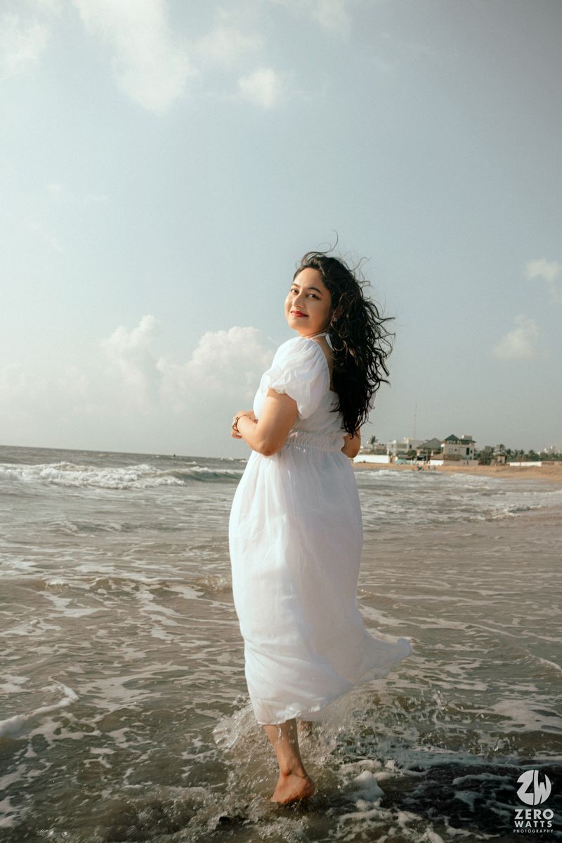 Woman in flowing white dress standing at edge of ocean waves on beach.