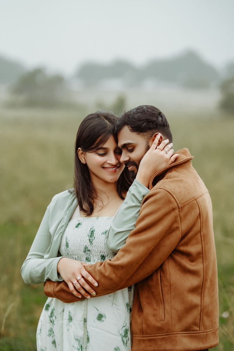 Romantic couple embraces in misty field, with woman in floral dress and man in brown jacket sharing tender moment.