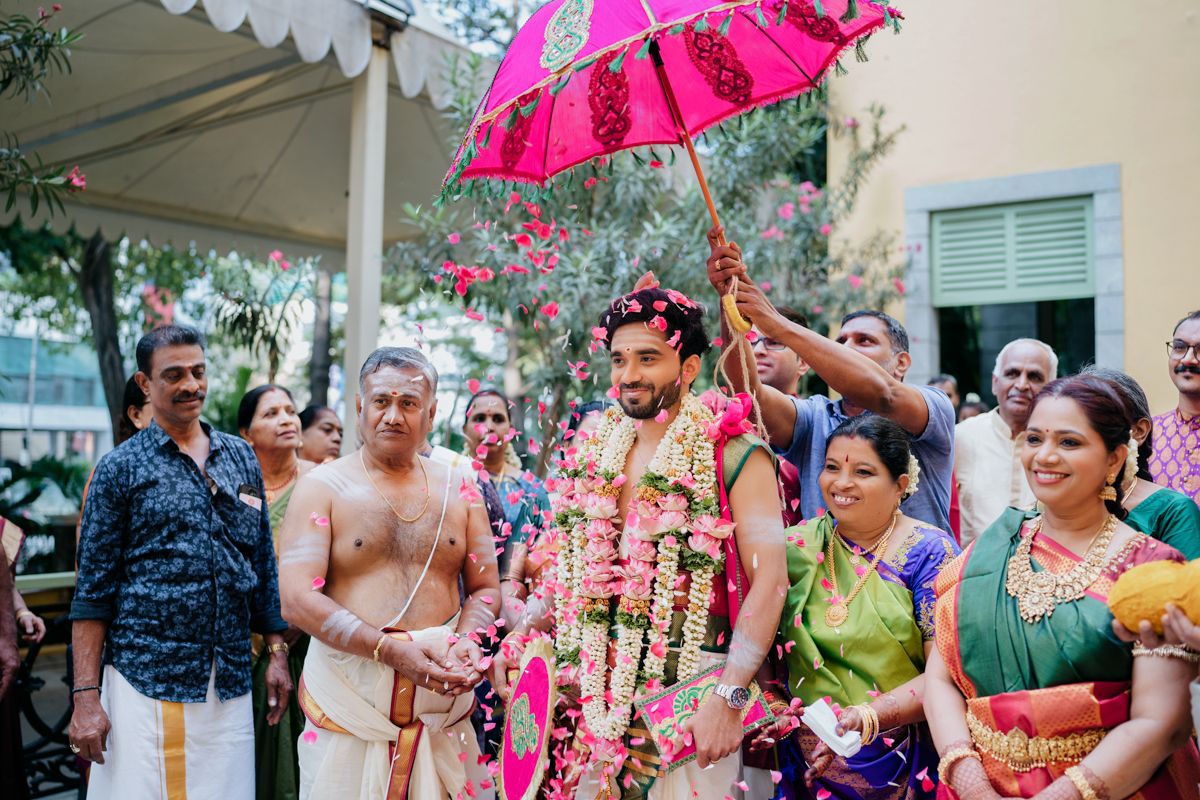 A traditional Indian wedding ceremony takes place under a pink umbrella with guests in colorful attire looking on.