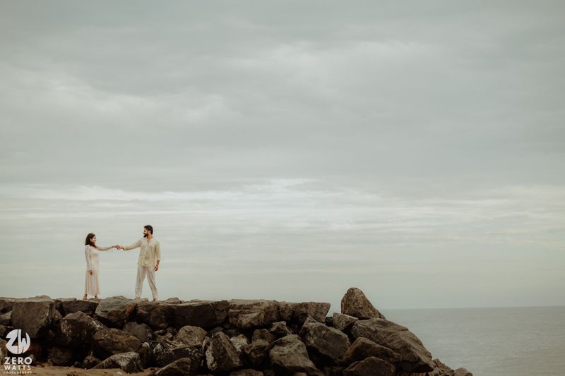 A romantic couple poses together on rocky coastal terrain against a misty gray sky.