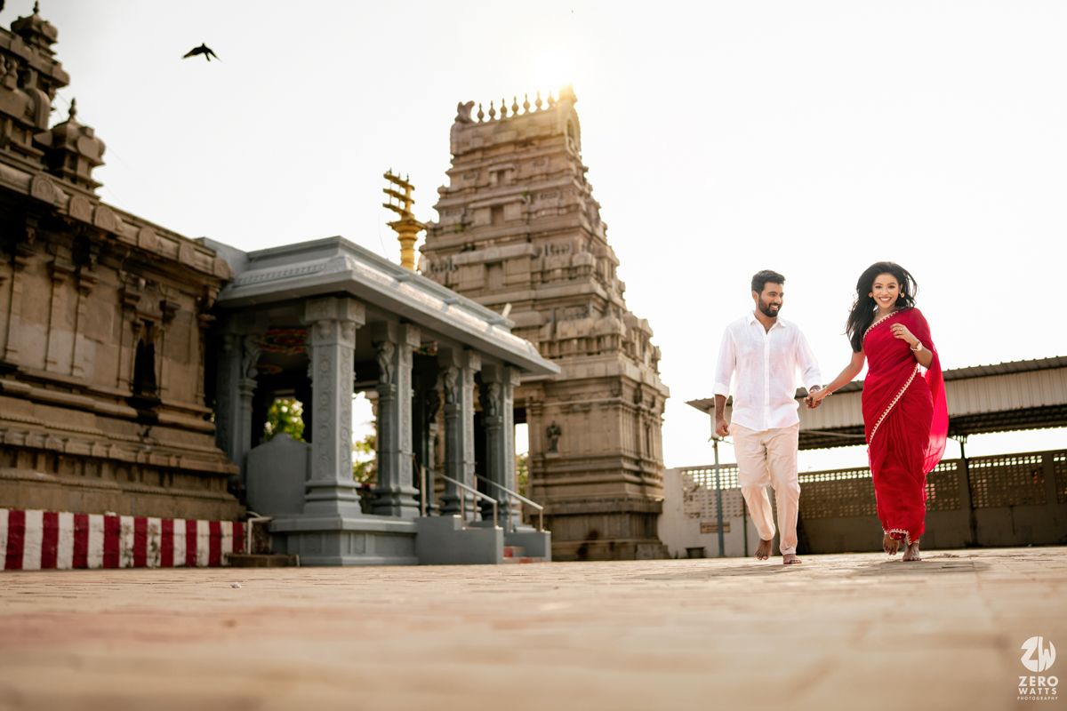 Two people walk together at a temple complex with ornate gopuram tower and traditional architecture in background.