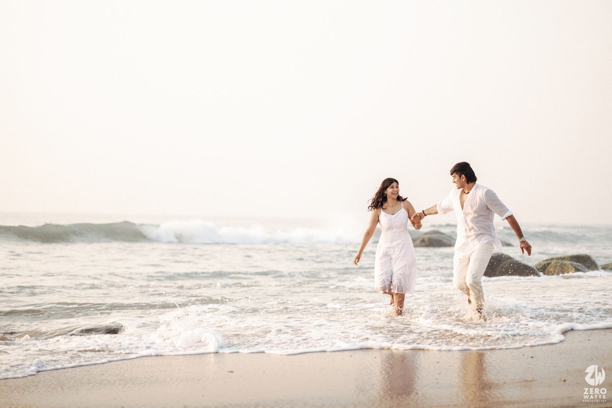 A couple in white clothing runs joyfully along the shoreline as ocean waves crash behind them during golden hour.