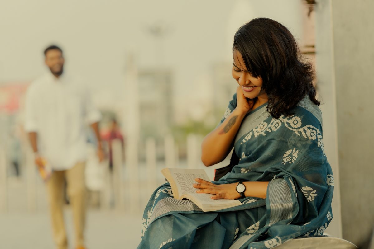 A person in a blue sari sits reading while talking on a mobile phone outdoors.