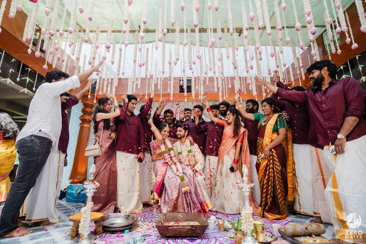 A festive Indian wedding ceremony with guests in maroon and white attire celebrating under hanging ribbons and decorations.