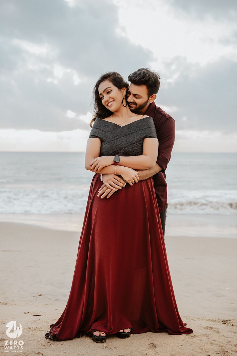 A romantic couple shares an intimate moment while dressed in black and burgundy on a cloudy beach at sunset.