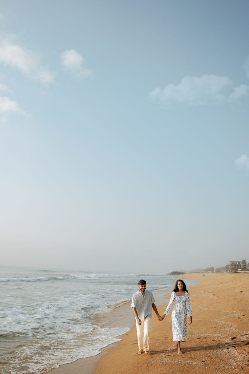 Couple walking hand in hand along an empty beach with waves crashing beside them.