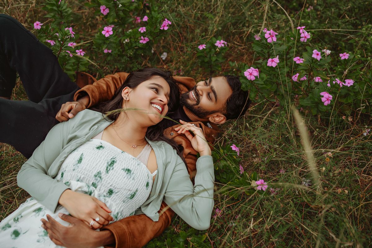 Couple lying in grass surrounded by pink flowers during romantic outdoor photoshoot.