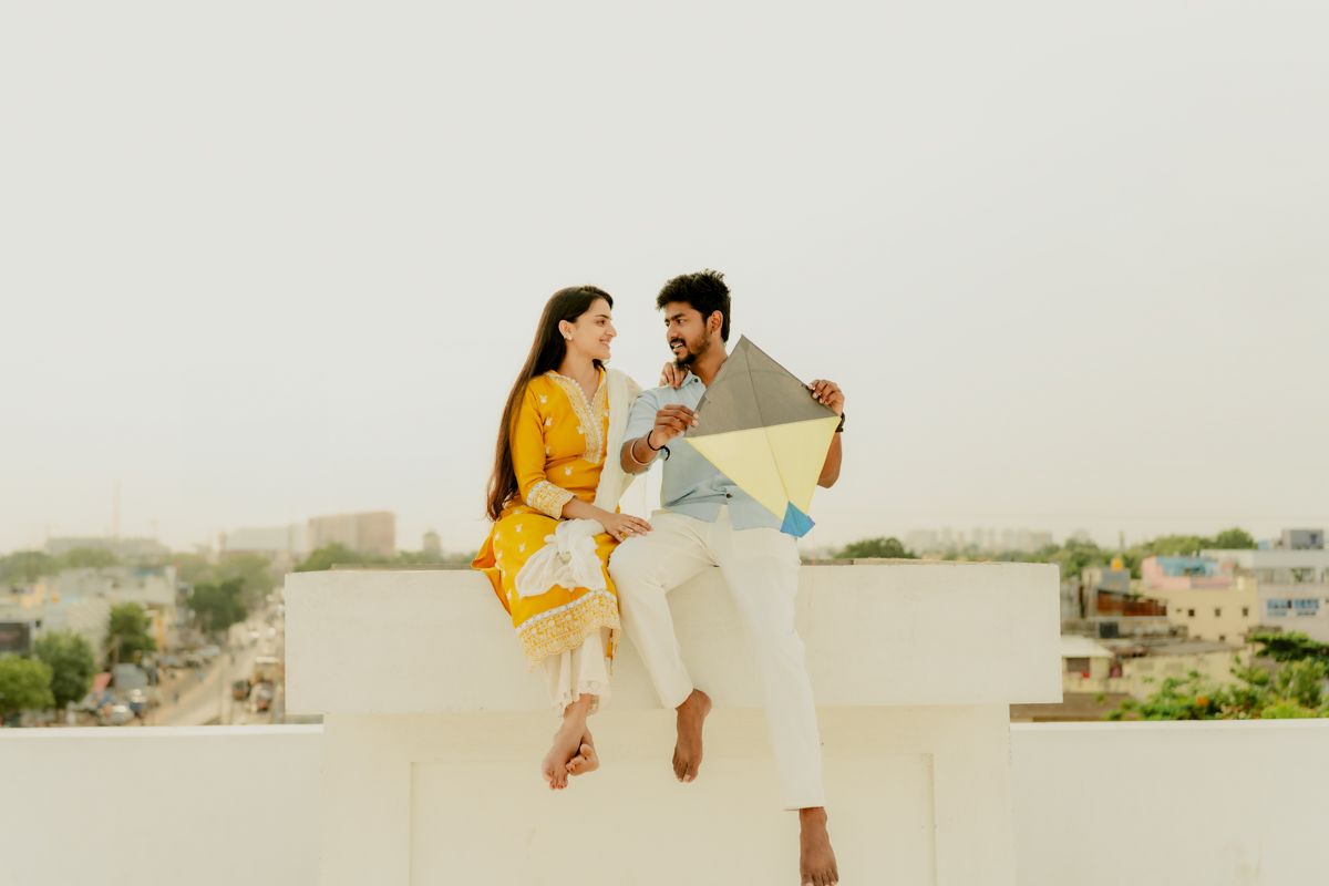 A couple sits together on a white ledge overlooking a cityscape while looking at something together.
