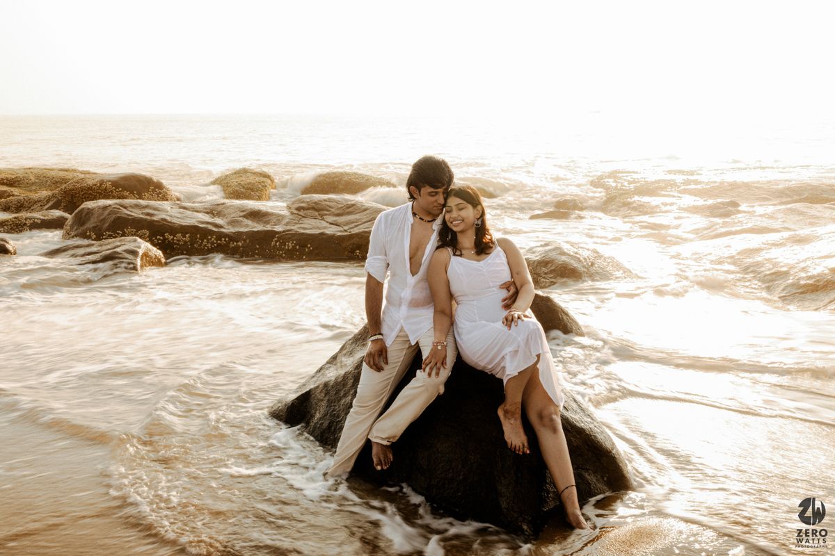 Couple in white clothing poses on rocky beach as golden sunlight bathes the scene.