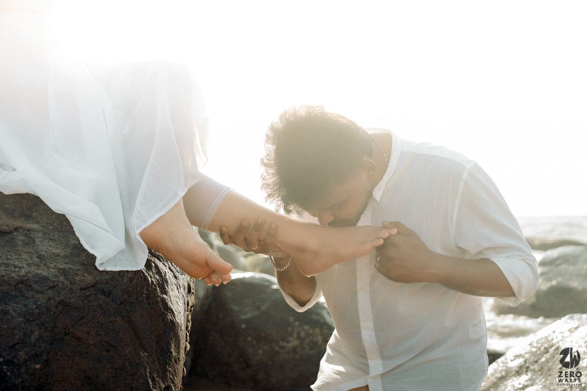 A couple in white attire sharing a romantic moment on coastal rocks during golden hour sunlight.