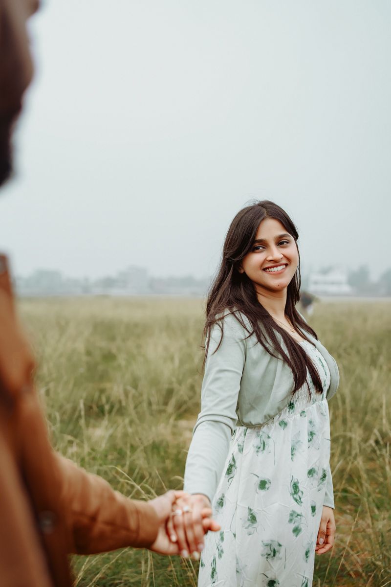 A person in a floral dress smiles while holding hands in a grassy meadow on an overcast day.