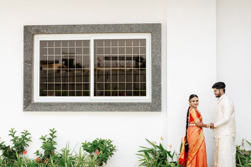 A couple in traditional Indian attire standing next to a window frame on a white wall with plants.