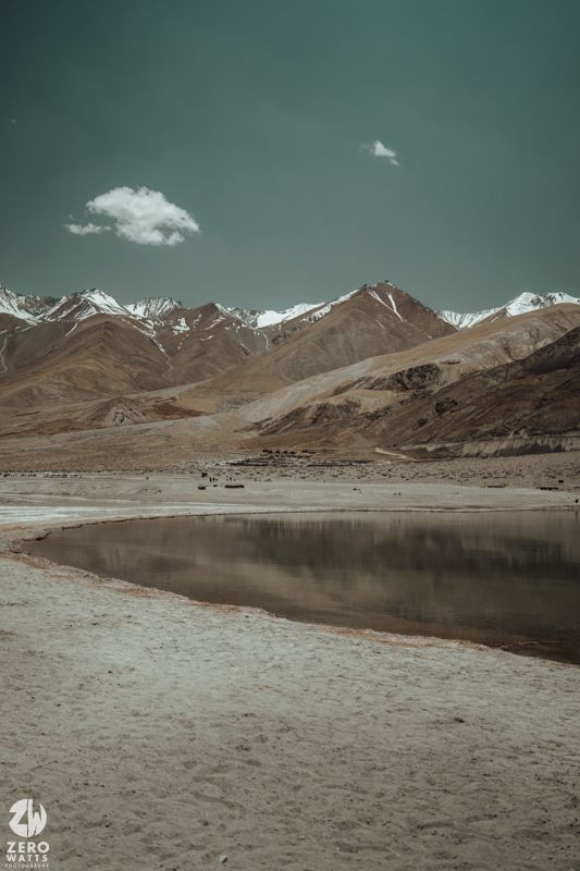 A serene mountain lake reflects brown peaks under a turquoise sky with scattered clouds.