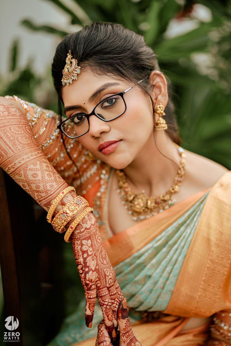 Indian bride in traditional attire wearing glasses, ornate gold jewelry and intricate henna designs on hands.