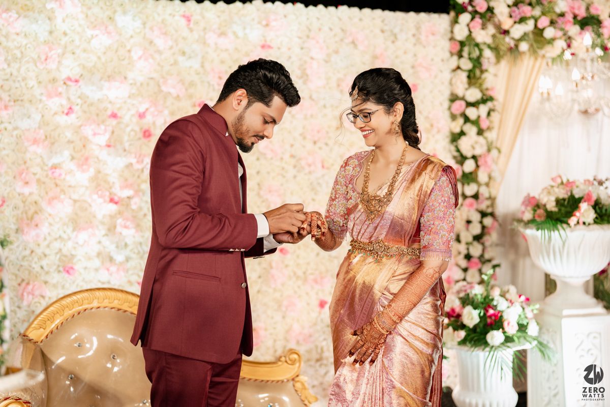 Couple in traditional attire performs wedding ceremony in front of floral backdrop at elegant ceremony venue.
