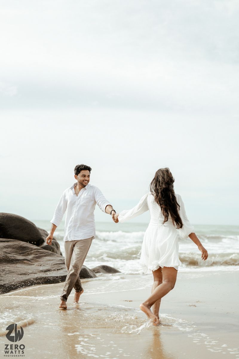 A romantic couple in white outfits holds hands while walking along a beach shore with crashing waves.