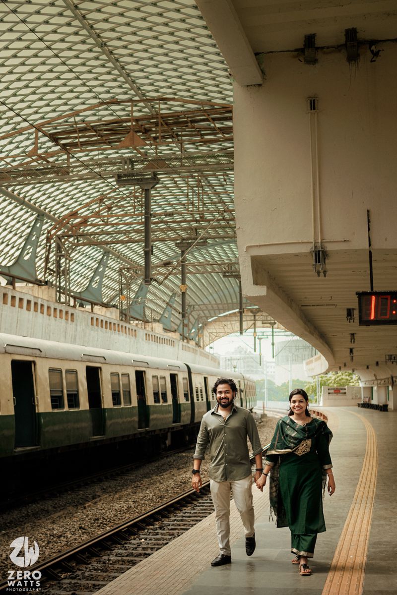 Couple walks hand in hand along a vintage railway platform with ornate architecture and natural lighting filtering through.