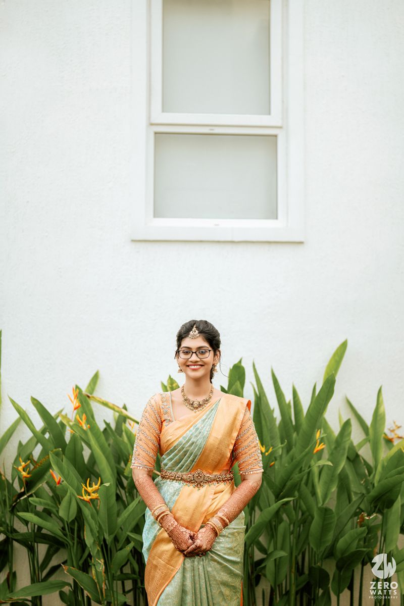 Person in a peach and mint silk saree poses elegantly against a white wall with tropical plants in the foreground.