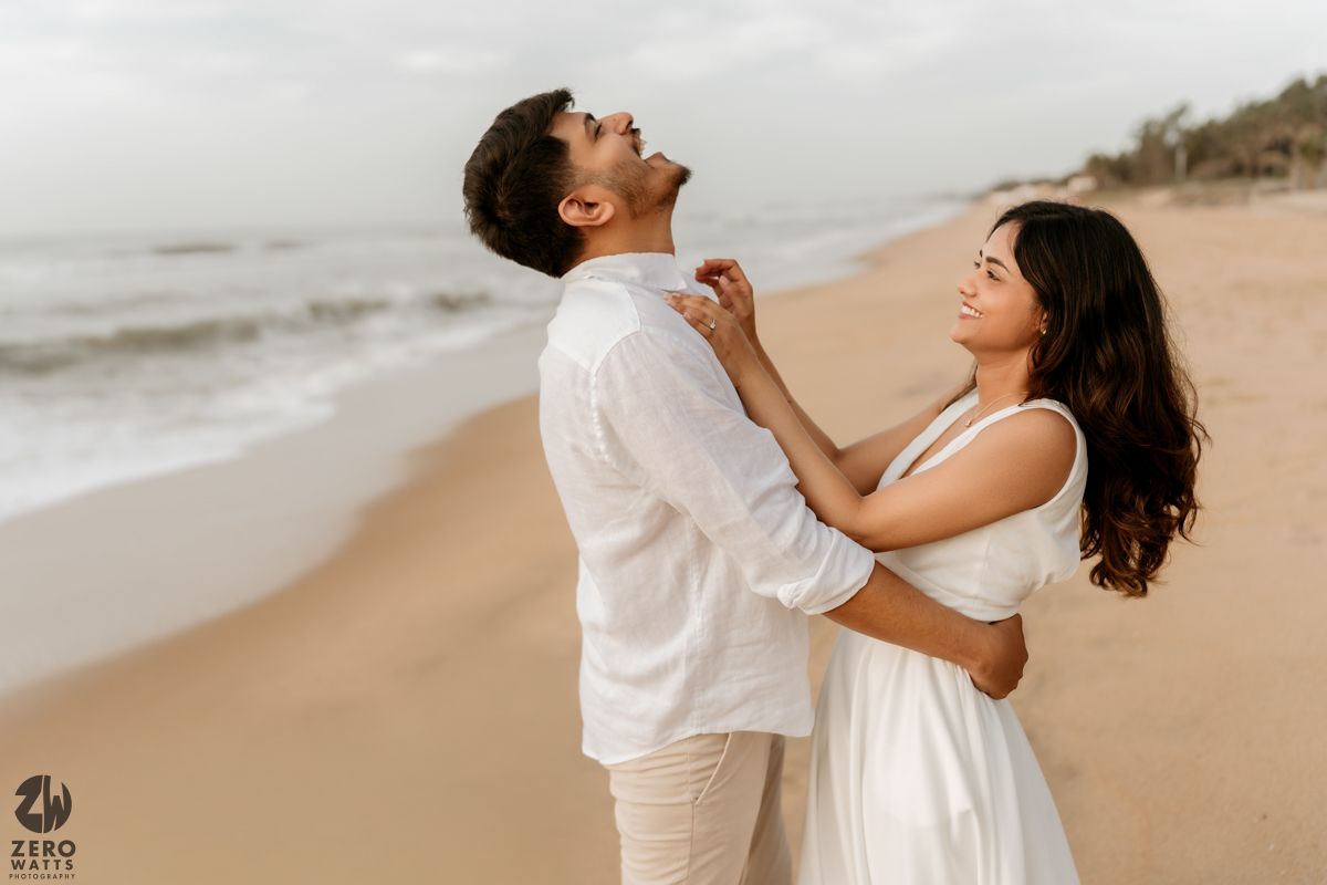 Playful couple in white outfits laugh and dance together on a cloudy beach.
