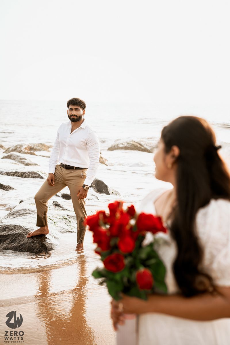 Couple poses on rocky beach with red roses against ocean backdrop.
