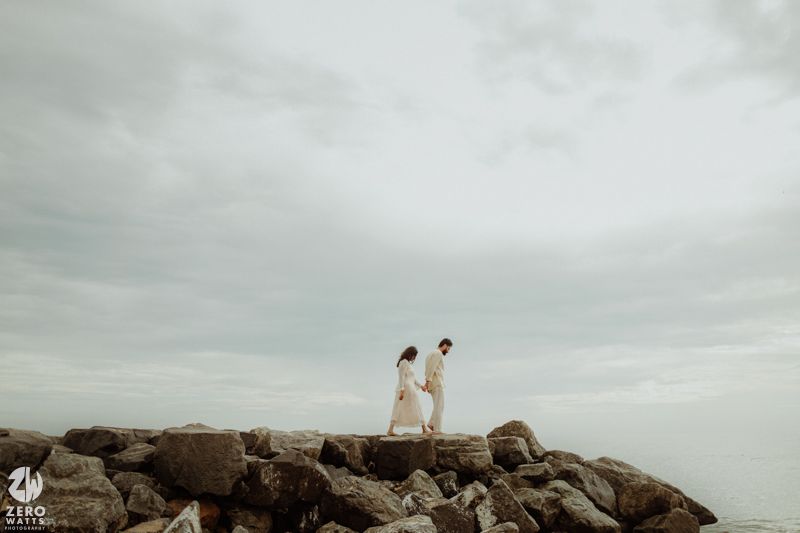 A couple in white attire standing on rocky coastal outcrop against cloudy sky.