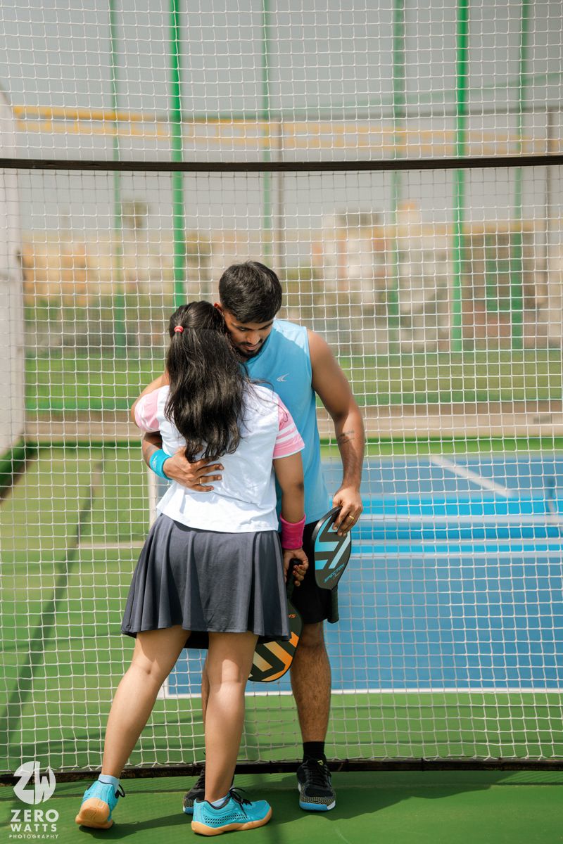 Couple in athletic wear embracing near white sports netting on a blue court.