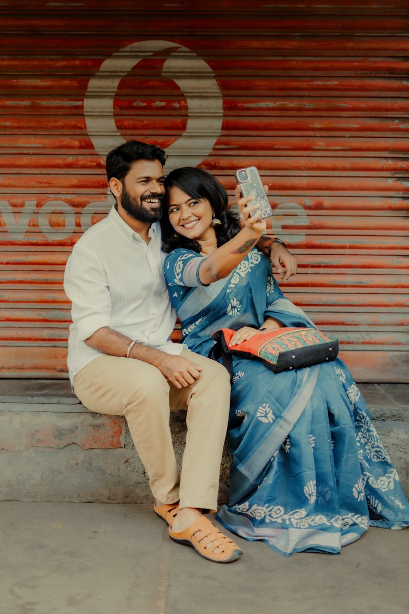 A couple in traditional and modern attire takes a selfie together against an orange shuttered wall.