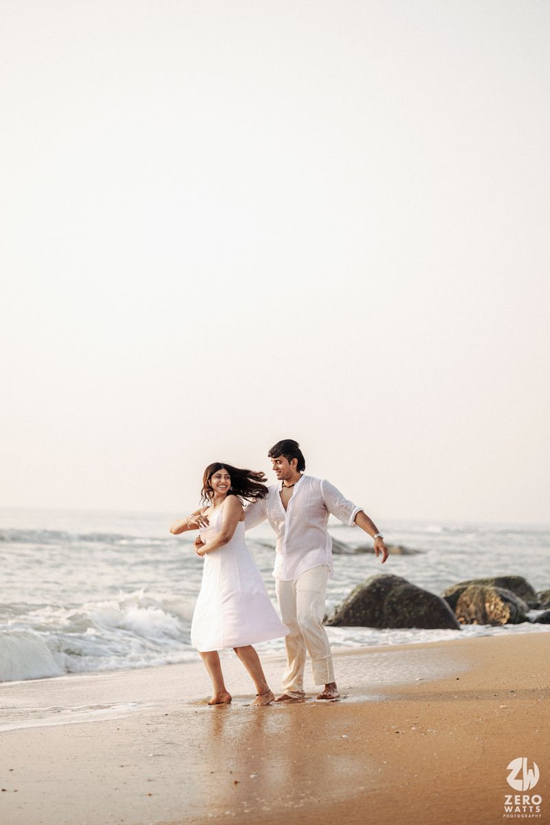 A couple in white outfits dances joyfully together on a beach at sunset with waves and rocks in the background.